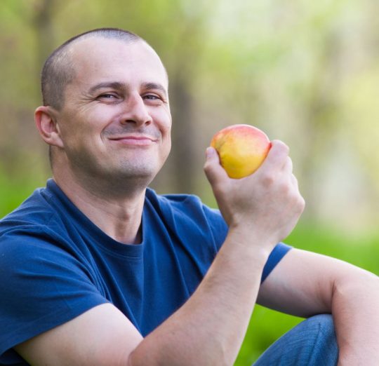 A smiling man in a blue T-shirt enjoying a fresh apple outdoors, surrounded by green nature.