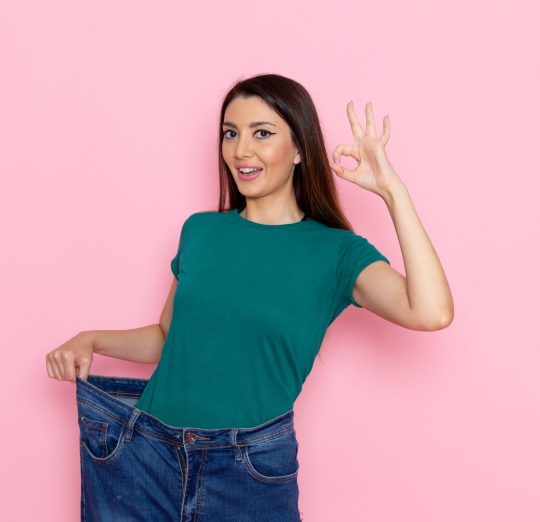 A happy woman in a green T-shirt showing off her weight loss by wearing oversized jeans, making an "OK" hand gesture against a pink background.