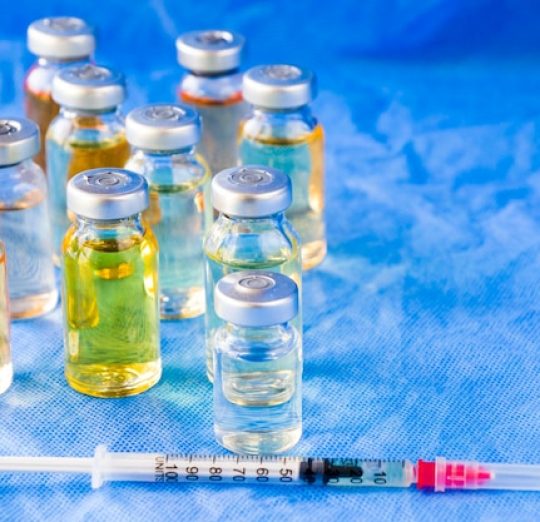 A set of medical vials containing liquid solutions with a syringe on a blue sterile background, symbolizing healthcare, vaccines, or medical treatments.